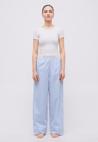 Γυναίκα όρθια, ξυπόλητη, φοράει λευκό εφαρμοστό μπλουζάκι και φαρδιά μπλε-λευκή ριγέ παντελόνα.