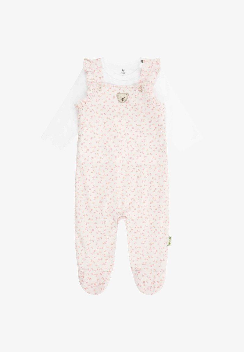 Helle pinke Baumwoll-Latzhose mit kleinem Blumenmuster, Rüschenträgern und Bär-Emblem. Unterhalb sichtbar ist ein weißes Langarmshirt.