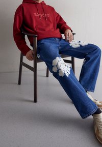 Bordowy hoodie z logo, niebieskie dżinsy z białymi wzorami tie-dye oraz beżowe sneakersy niskie, wszystko noszone podczas siedzenia na drewnianym krześle.
