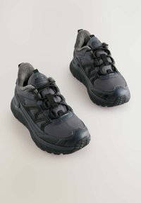 Par af mørkegrå og sorte sneakers med tykke såler, snørebånd i løkker og blød indersål, fotograferet på en lys baggrund.