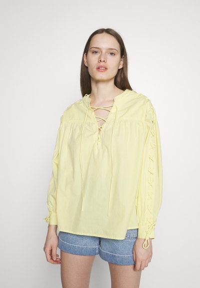 Blouse jaune à manches longues avec un décolleté lacé et des accents de lien sur les manches, fabriquée en tissu léger. Associée à un short en denim.