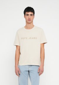 Noor mees lühikeste tumejuustega, kandes beeži värvi "PEPE JEANS" T-särki ja hele siniseid teksapükse, seismas ühevärvilise valge tausta ees.