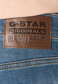 Ruskea nahkalätkä sinisellä denim-kankaalla, jossa kohopainettu teksti "G-STAR ORIGINALS RAW DENIM." Lätkässä näkyy ompeleet ja sileä pinta.