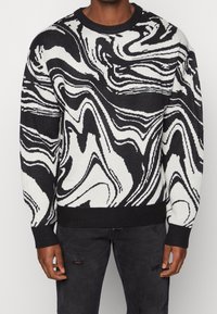 Pull à manches longues noir et blanc avec motif abstrait marbré porté par une personne portant un jean sombre usé.