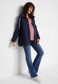 Felpa con zip colore navy, con cappuccio e stringhe bianche, abbinata a una maglietta a righe rossa e bianca e jeans a zampa d'elefante blu, completata da sneakers bianche.