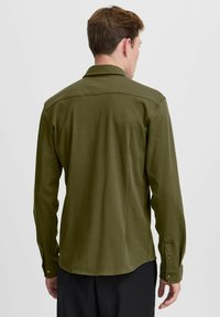 Olive green long-sleeve shirt in a smooth fabric; features a collared neck, button cuffs, and a slightly curved hemline.