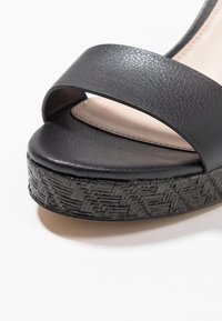 Černé sandály s širokým, hladkým syntetickým páskem a strukturovanou klínovou podešví s tkaným vzorem. Jemné béžové lemování na stélce.