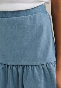 Šviesiai mėlyna džinsinė sijonas su pakopiniu raukšlėtu dizainu, pasižymintis lygia tekstūra ir subtiliu įstrižiniu audimo raštu. Matomi elastingo juosmens detalės.