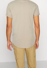 Bežové tričko s krátkými ohrnutými rukávy, kulatým výstřihem a delším zadním lemem, vyrobené z měkké směsi bavlny. Žádné viditelné vzory.