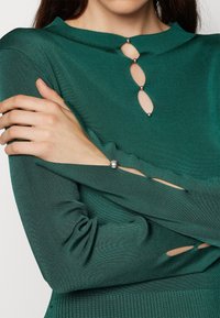 Haut en maille côtelée verte avec un col montant, une ouverture en forme de goutte d'eau ornée de boutons décoratifs, et des manches fendues avec des accents métalliques.