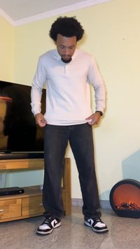 Camisa de manga larga beige claro con cuello, jeans negros y zapatillas en blanco y negro. De pie en el interior, con las manos ajustando el dobladillo de la camisa.