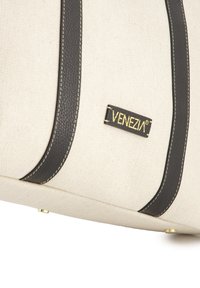 Torba płócienna w kolorze off-white z czarnymi skórzanymi akcentami, z podwójnymi uchwytami, złotą metką z logo, białymi przeszyciami i metalowymi nóżkami dla stabilności.