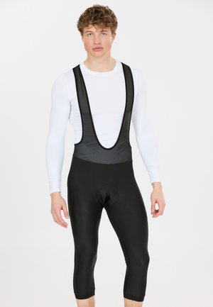 Nuori mies, jolla on mustat pyöräilyhenkselit ja valkoinen pitkähihaisen kompressiopaita, seisoo yksinkertaisen valkoisen taustan edessä.