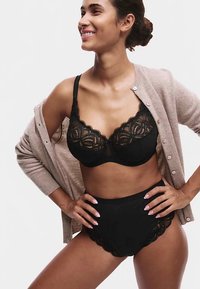 Sort blonderetop med bølget kant kombineret med højtaljede sorte trusser med blondekant. Modellen har en tanfarvet cardigan på. Glat stoftekstur.