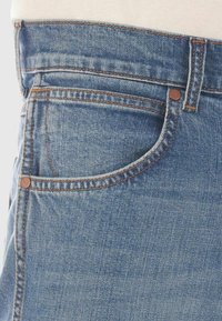 Acercamiento a un bolsillo de jeans de mezclilla azul con remaches de cobre visibles y costura naranja sobre un fondo blanco.
