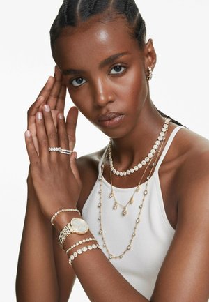 Frau trägt weißes Tanktop mit Gold- und Diamantschmuck, darunter mehrlagige Halsketten, Armbänder, Ringe, Ohrringe und eine goldene Uhr.