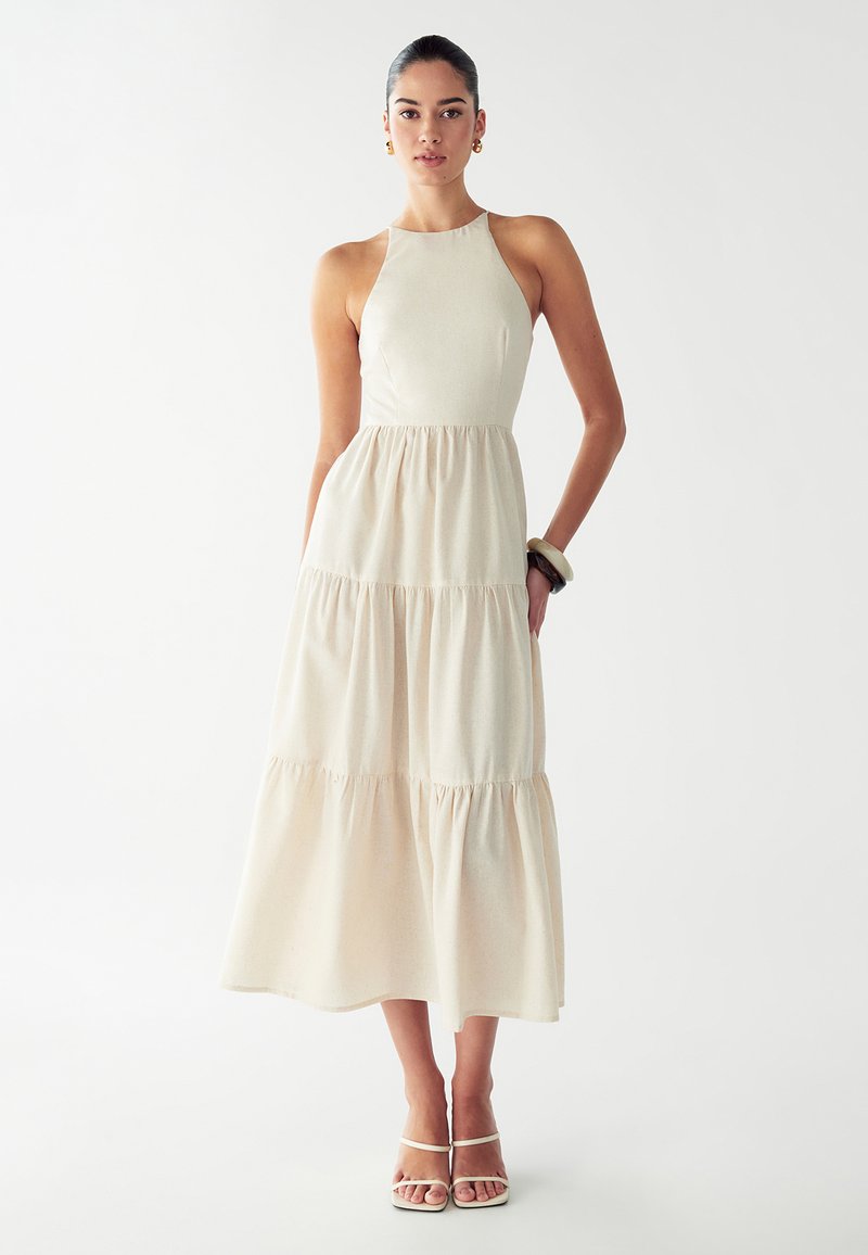 Nainen, joka on pukeutunut hihattomaan, kerrokselliseen beigen midi-mekkoon, jossa on halterneck-olkaimet, yhdistäneenä paljettikäsiin ja valkoisiin nilkkaremmikenkiin, seisoo valkoista taustaa vasten.