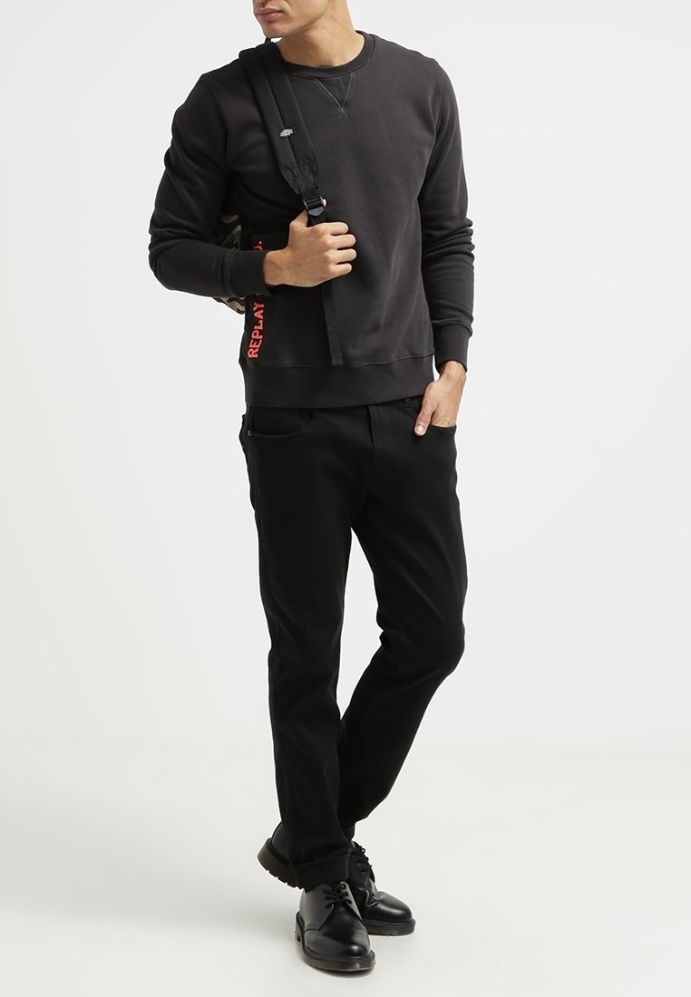 Svart sweatshirt med rund halsringning och ribbade muddars, ihop med svarta byxor. Svarta skor och en axelremsväska med röd text "REPLAY".