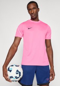 Ροζ αθλητική μπλούζα Nike με κοντά μανίκια, στρογγυλή λαιμόκοψη και μαύρο λογότυπο. Σκούρες μπλε σορτς με ροζ λογότυπο. Κρατάει μια λευκή μπάλα ποδοσφαίρου.