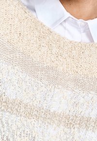 Vaaleanbeige neulottu villapaita, jossa on teksturoidut kuviot ja yhdistelmä avoimia virkkauskuvioita sekä kiinteää neulosta. Valkoinen kauluksellinen paita näkyy sen alla.