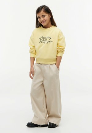 Niña sonriente está de pie con las manos en los bolsillos, lleva una sudadera amarilla "Tommy Hilfiger", pantalones anchos beige y zapatos negros sobre fondo blanco.