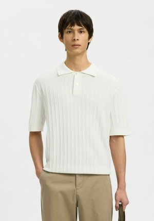 Jeune homme portant un polo en maille blanc à manches courtes et un pantalon beige, debout, une main dans la poche et tenant un sac.