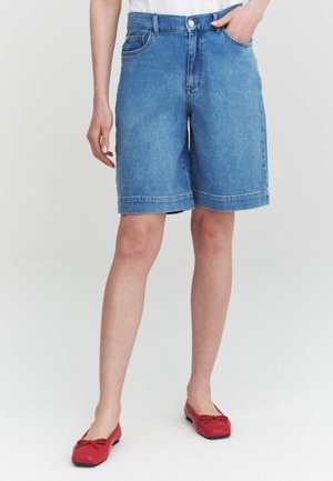 Person trägt mittelblaue Jeansshorts mit Vordertaschen und roten Ballerinas und steht vor einem einfarbigen hellen Hintergrund.