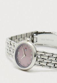 Sudraba metāla rokas pulkstenis ar rozā ciparnīcu, kristāla stundu atzīmēm, romiešu cipariem pie 6 un 12, kā arī logo tuvumā 12 stundu pozīcijai.