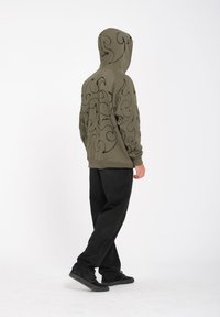 Hoodie verde oliva con motivo a spirali nere, la texture del tessuto appare morbida. Abbinato a pantaloni neri e scarpe scure, modello di spalle.