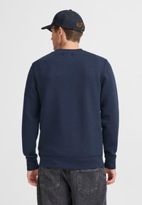 Hombre vistiendo un sudadera azul marino, gorra negra y jeans negros desgastados, de pie de espaldas frente a un fondo blanco liso.