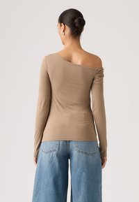 Μπλούζα με μακριά μανίκια και έξω τους ώμους σε ανοιχτό καφέ ύφασμα με εφαρμοστή σιλουέτα. Διαθέτει ραβδώσεις στους ώμους. Συνδυάζεται με ανοιχτό μπλε τζιν παντελόνι με φαρδιά πόδια.