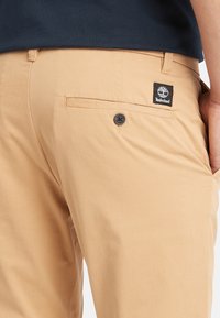 Pantaloni beige con una texture liscia, dotati di una tasca posteriore con un bottone nero e un'etichetta nera del logo Timberland sopra la tasca.