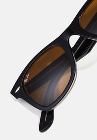 Black sunglasses with brown lenses, rectangular frames, and a glossy finish. Features metal accents and Ray-Ban branding on the lens.