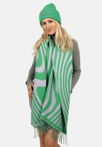 Grüne gestrickte Mütze und übergroßer Schal mit grünen und grauen wellenförmigen Mustern. Der Schal hat fransige Kanten und eine weiche Textur.