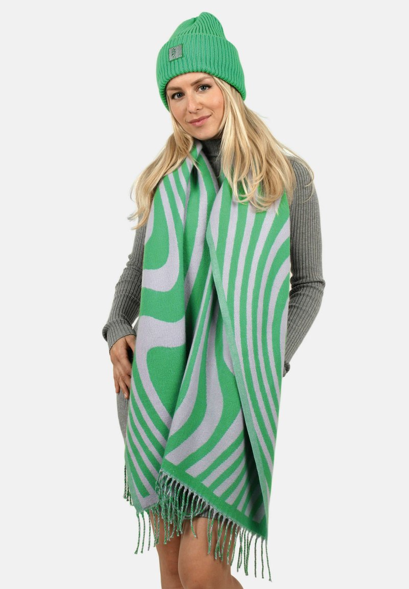 Grüne gestrickte Mütze und übergroßer Schal mit grünen und grauen wellenförmigen Mustern. Der Schal hat fransige Kanten und eine weiche Textur.