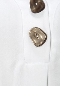 Weißer Stoff mit strukturierter Oberfläche, versehen mit zwei unregelmäßigen, hellbraunen Knöpfen mit glänzender Oberfläche und einem zentralen Loch zum Befestigen.