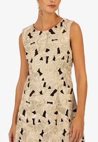 Beige bloemenbroderie jurk met uitsnijdingen, voorzien van een mouwloos ontwerp en een gladde textuur. Opvallende zwarte accenten verbeteren het patroon.