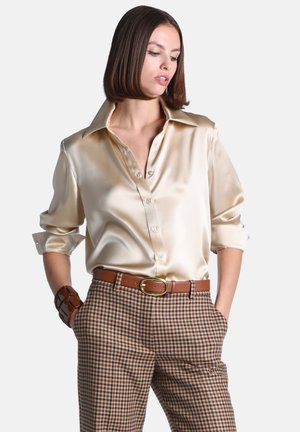 Femme portant un chemisier en soie crème brillant avec manches retroussées, pantalon à carreaux marron, ceinture marron et large bracelet marron, mains dans les poches.