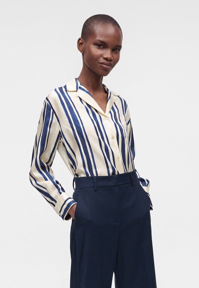 Femme aux cheveux courts portant une chemise à rayures verticales crème et bleues rentrée dans un pantalon large bleu marine, les mains dans les poches.