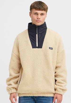 Beige fleece pullover with a high collar, featuring a navy insert, half zipper, and textured surface. Detailed with a small logo on the chest.