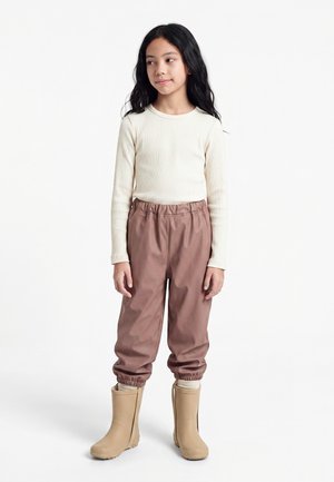 Enfant aux longs cheveux foncés portant un pull à côtes crème à manches longues, un pantalon mauve, des bottes de pluie beiges, debout sur un fond blanc.