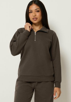 Jeune femme aux longs cheveux foncés portant un sweat zippé marron foncé et un pantalon assorti, tenant la tirette de la fermeture éclair près de son cou.