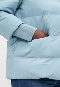 Chaqueta acolchada de color azul claro con puños acanalados, que presenta un bolsillo lateral y un diseño acolchado para el aislamiento. Material exterior suave y brillante.