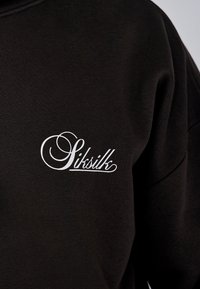 Svart sweatshirt med en slät yta och en vit kursiv logotyp på vänster bröstområde. Enkel, minimalistisk design.