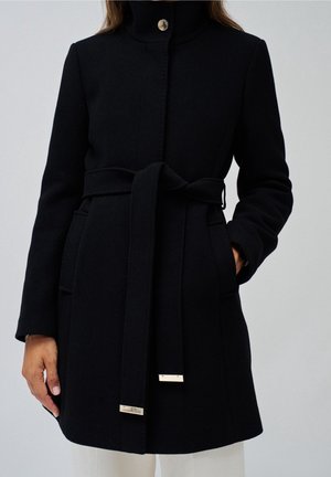 Black belted wool coat with high collar, side pockets, and silver metal accents on the belt ends, worn over white pants.