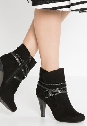 Czarne zamszowe botki na wysokim obcasie z ozdobnymi łańcuszkami i paskami, noszone z czarno-białą spódnicą w paski.