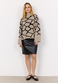 Beiger Pullover mit schwarzem abstraktem Muster, hohem Kragen, weiten Ärmeln; kombiniert mit einem schwarzen Leder-Bleistiftrock mit strukturierter Oberfläche. Schwarze Schuhe.