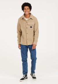Giacca in pile marrone chiaro con bottoni, una tasca frontale e patch con logo, abbinata a jeans di denim blu e sneaker nere. Texture liscia in tutto.
