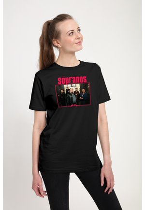 Kvinde iført sort T-shirt med et gruppefoto og rød tekst "The Sopranos" ser let opad mod højre.