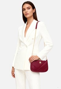 Borsa a tracolla in pelle color bordeaux con motivo trapuntato, dettagli in oro e tracolla a catena, tenuta da una persona che indossa un blazer bianco e pantaloni.